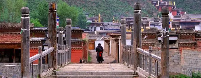 Timber posts and stone steps on the walkway into a Langmusi monastery, a figure in red heading in