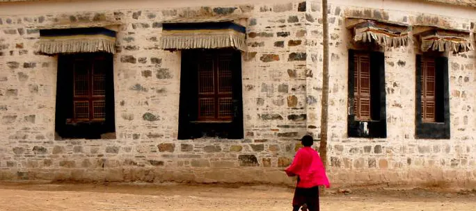 A lane in Langmusi — stone walls, black window frames, pleated valances above each window, a figure in red passing