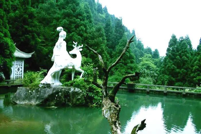 麻姑仙境：一尊白色雕像立在池中的石头上，旁边是一只鹿，池水是深绿色，四周全是树