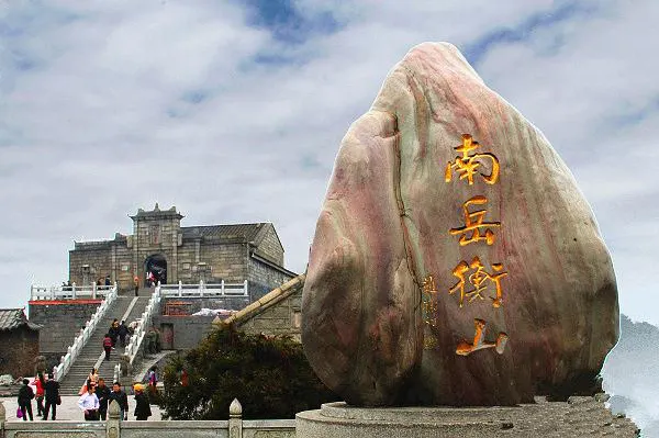 峰顶的“南岳衡山”石刻：一块粉褐色巨石立在台座上，四个金字竖排，后面是石阶和门楼，游客在台阶上上下下