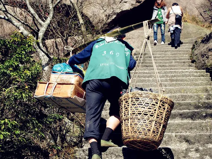 A porter with a bamboo pole, cardboard boxes on one end and a woven basket on the other, on the same steps as everyone else
