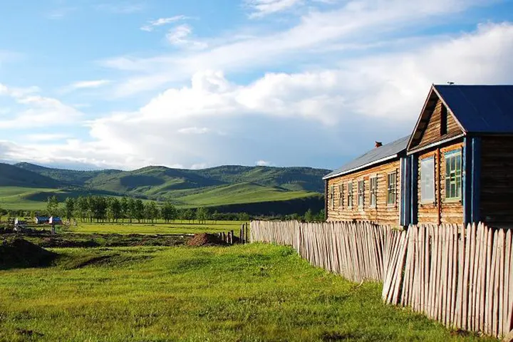 A log house behind a fence of split poles, grass slopes stacked up behind it