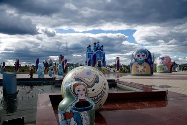 The matryoshka square — painted dolls the height of a house, castle fronts behind, storm cloud overhead