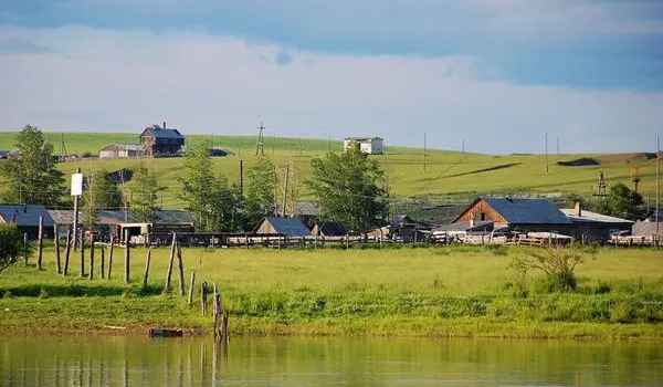The village from the water — log walls, pole fences, a grass slope rising behind the roofs