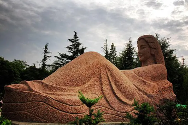 A reclining Buddha carved in stone on a lawn, pines behind it
