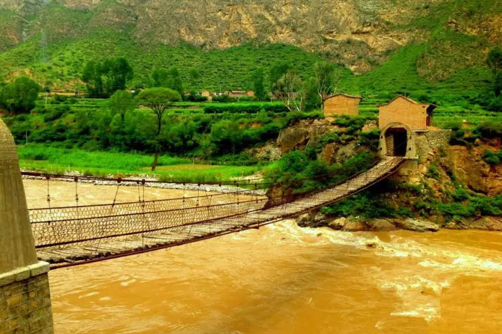 A chain suspension bridge over the silty Yellow River, with loess hills and a brick gateway on the far bank