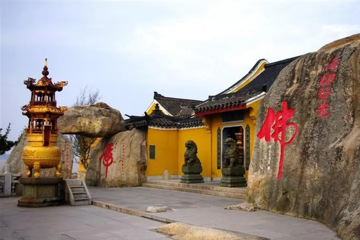 A temple gate tucked under an overhanging boulder, a bronze burner to the left and a red Buddha character cut into the rock