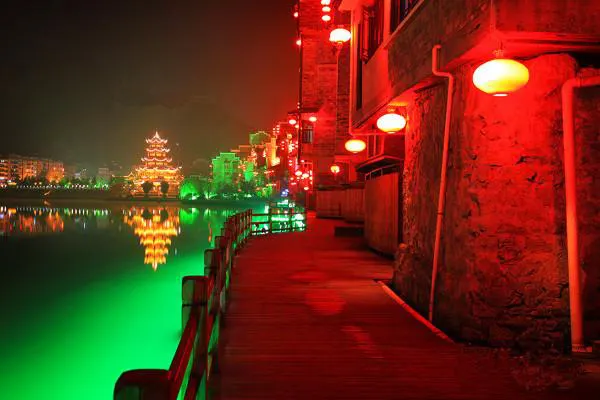 The riverside walk after dark: lanterns along the railing, a lit pavilion across the water